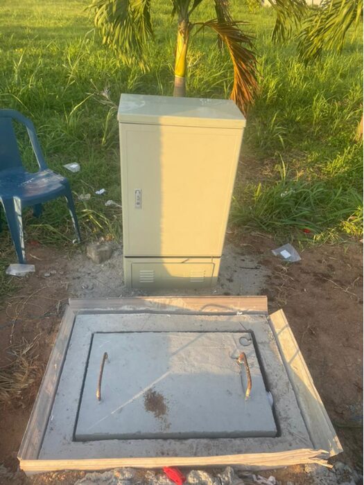 Closed outdoor fibre cabinet standing on a concrete plinth behind a freshly concreted access chamber with metal lifting handles